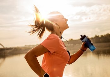 Eine Frau joggt laechelnd mit Wasserflasche in der Hand an einem See die Sonne geht unter