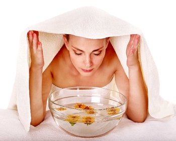 Eine Frau macht ein Dampfbad fürs Gesicht, indem sie sich über eine Wasserschüssel mit Blumen beugt und ein Handtuch über den Kopf hält