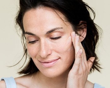 Brünette Frau mit geschlossenen Augen, die Creme auf ihre linke Wange aufträgt