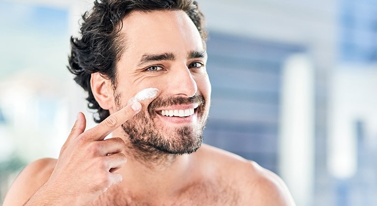 Junger Mann mit Drei-Tage-Bart trägt Gesichtspflege auf seine rechte Wange auf