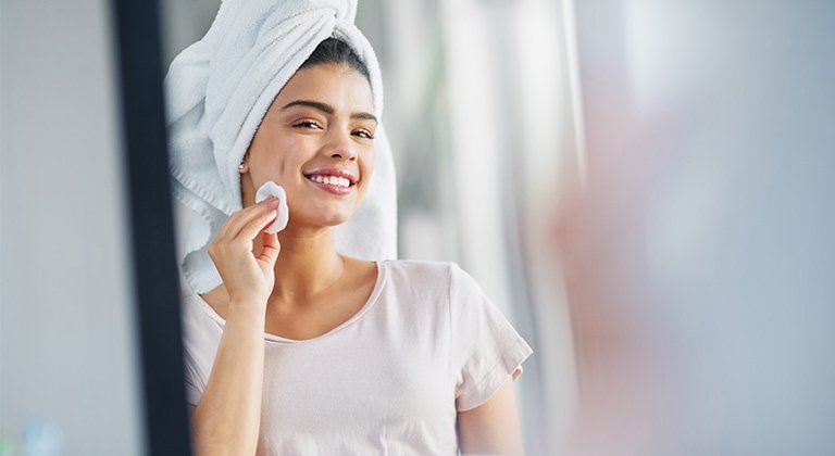 Lächelndes Gesicht einer Frau mit einem Wattepad in der Hand und einem Handtuch auf dem Kopf