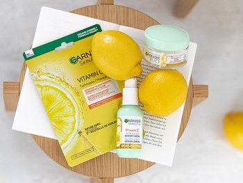 Garnier Vitamin C Produktset mit Tuchmaske, Serum-Creme, Cremetiegel und Zitronen auf Holzstuhl.