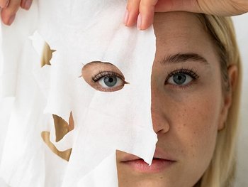 Blonde Frau, die sich eine Tuchmaske vor ihre rechte Gesichtshälfte hält