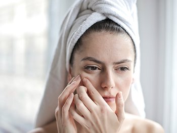 Eine junge Frau, hat ihre Haare mit einem Handtuch eingewickelt und fühlt mit der Hand Hautunreinheiten auf ihrer rechten Wange