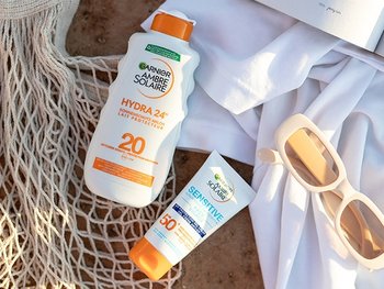 Zwei Ambre Solaire Sonnenschutzprodukte im Sand neben einer Sonnenbrille und einem aufgeschlagenem Buch