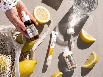 Hand einer Frau, die das Vitamin C Nachtserum über einem Boden mit Zitronen und weiteren Vitamin C Produkten hält