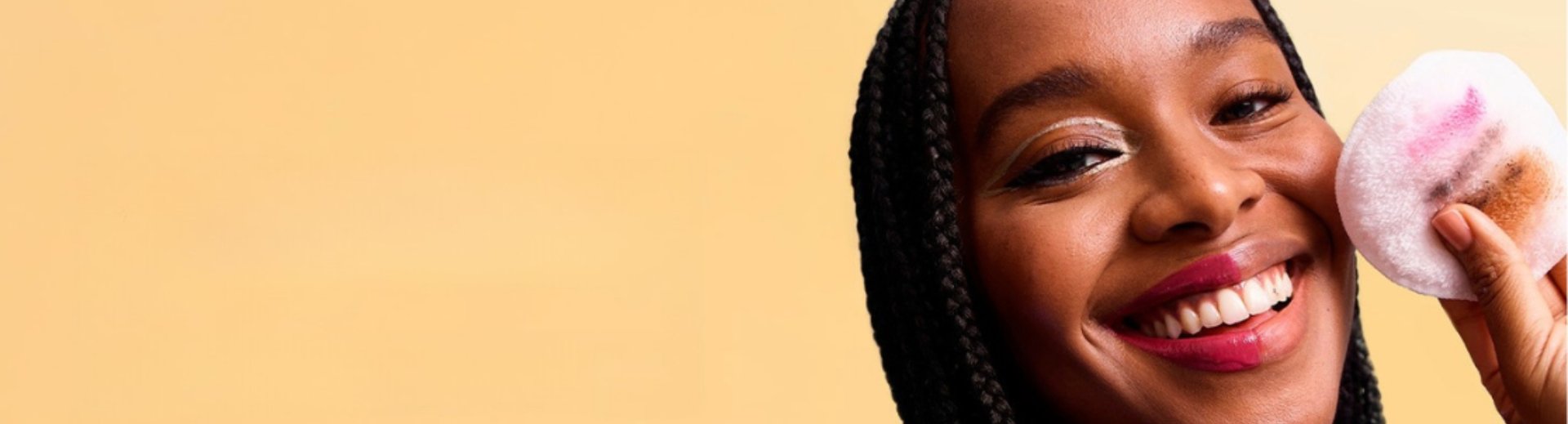 Lächelnde Frau vor gelbem Hintergrund schminkt ihr Make-up mit Mizellenwasser ab und hält ihr Wattepad in die Kamera