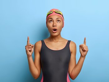 Frau im Badeanzug vor einer blauen Wand, die mit ihren Zeigefingern nach oben zeigt