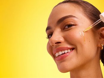 Junge, lächelnde Frau trägt ein Serum mit Vitamin C Derivaten auf die Wange auf