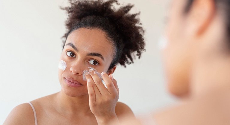 Eine junge Frau, vor einem Spiegel stehend, cremt sich ihr Gesicht ein
