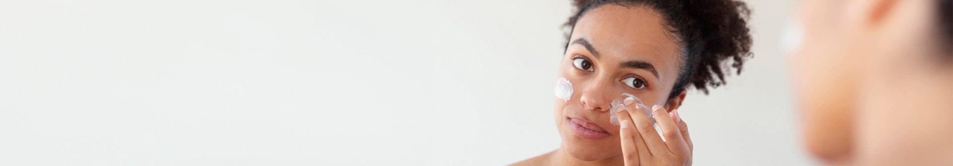 Eine junge Frau, vor einem Spiegel stehend, cremt sich ihr Gesicht ein