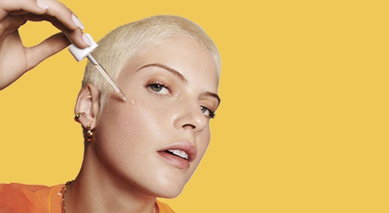 Ein junge Frau mit blonden kurzen Haaren trägt auf ihre Wange Vitamin C Serum mit Pinzette auf