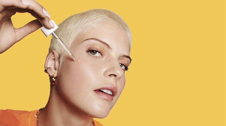 Ein junge Frau mit blonden kurzen Haaren trägt auf ihre Wange Vitamin C Serum mit Pinzette auf