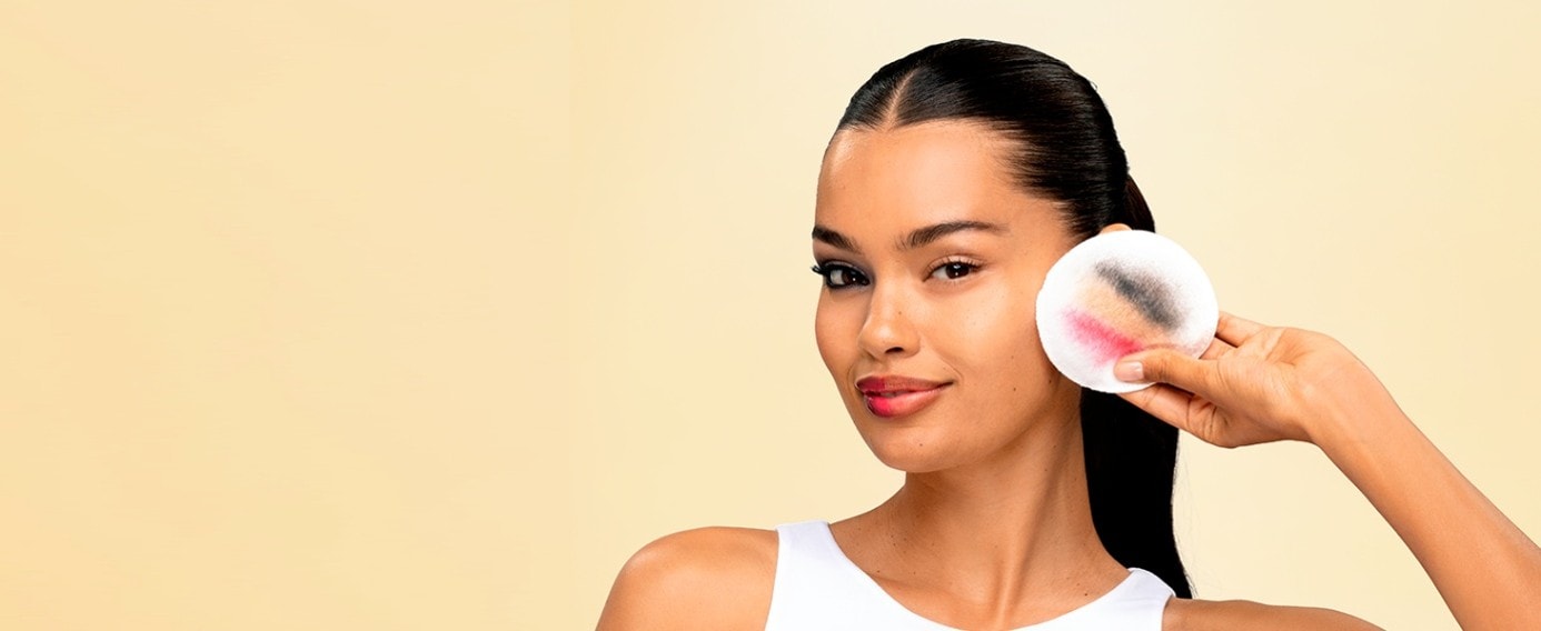 Eine junge Frau hält ein Wattepad mit Make-Up-Rückständen darauf. Ihr rechts Auge ist komplett ungeschminkt.