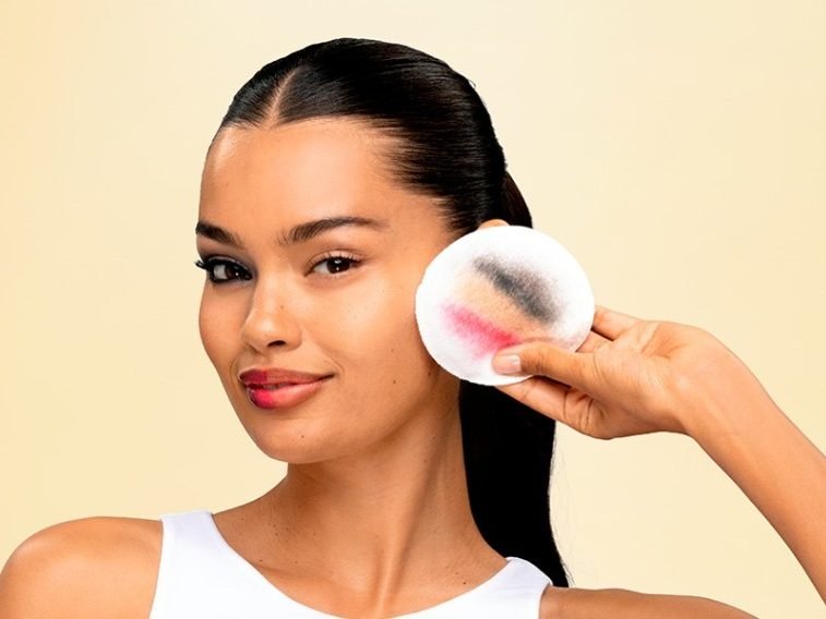 Eine junge Frau hält ein Wattepad mit Make-Up-Rückständen darauf. Ihr rechts Auge ist komplett ungeschminkt.