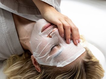 Blonde Frau mit einer Tuchmaske im Gesicht, die sich eine Hand vor das Gesicht hält