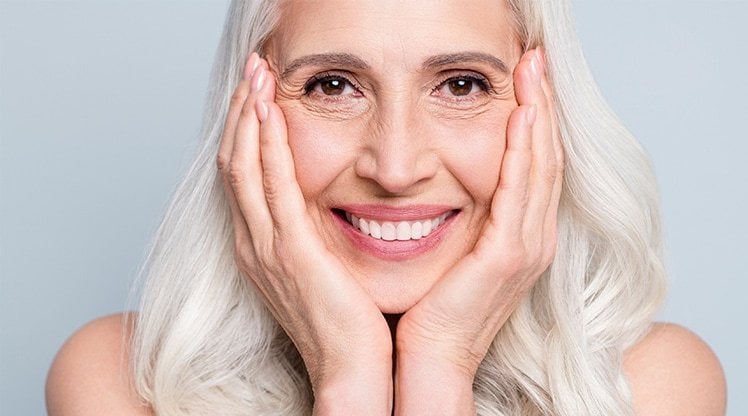 Frau mit langen grauen Haaren, lächelnd, ihr Gesicht auf ihre Hände gestützt