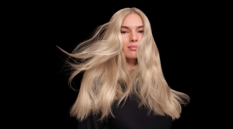 Frau steht vor schwarzem Hintergrund, ihr langes, blondes Haar weht zur Seite
