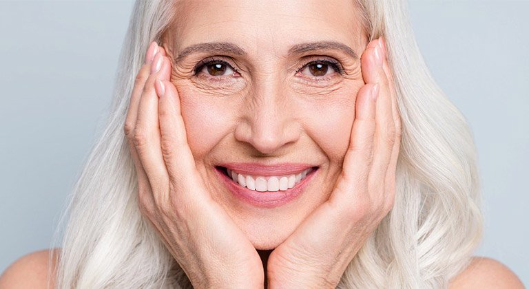 Lächelnde ältere Frau mit langem weißlich grauem Haar, die ihr Gesicht auf ihre Hände stützt