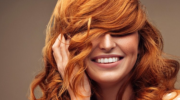 Lächelnde Frau hält sich ihre langen, roten Haare vor die Augen