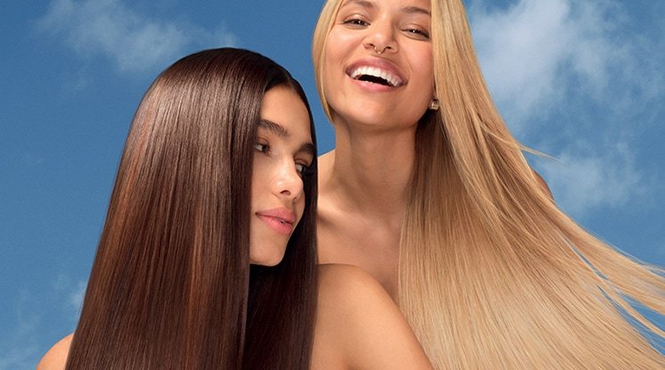 Zwei lächelnde junge Frauen mit langen Haaren vor blauen Himmel