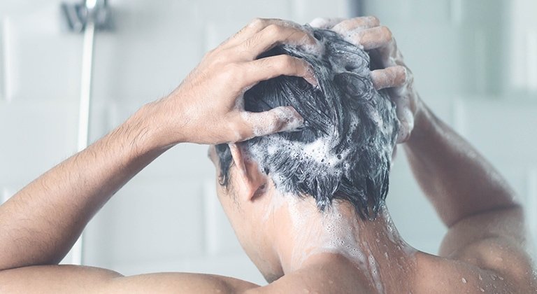 Eine Person steht unter der Dusche und massiert Shampoo in ihre nassen Haare ein, während Wasser herunterläuft