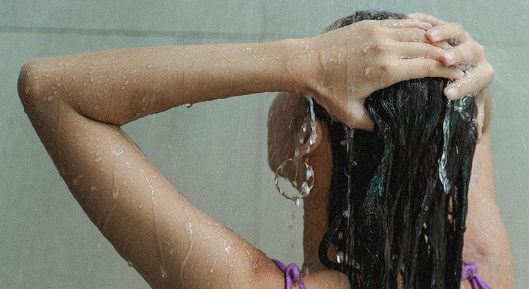 Eine junge Frau steht unter der Dusche und streift sich mit ihren Händen durch ihre langen Haare