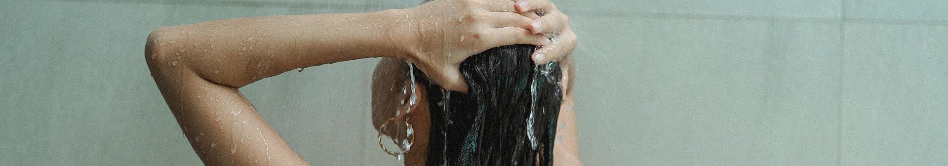 Eine junge Frau steht unter der Dusche und streift sich mit ihren Händen durch ihre langen Haare