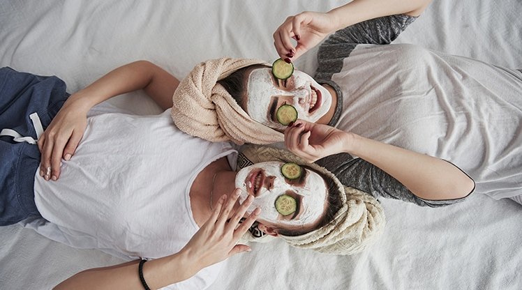 Zwei Frauen mit Gesichtmasken mit Gurkescheiben liegen auf einem Bett