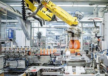 Eine Maschine mit gelbem Greifarm verschiebt Shampoo-Verpackungen auf einem Fließband