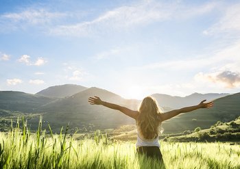Junge Frau mit dem Rücken zugewandt, auf einer Wiese stehend, grüßt die Arme ausbreitend die Sonne
