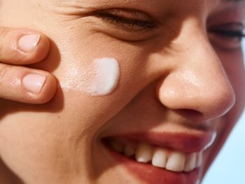 Gesicht einer Frau von der Seite, die lächelnd mit zwei Fingern Sonnencreme auf ihr Gesicht aufträgt