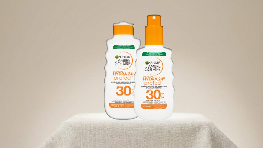 Blonde Frau, die die Garnier Ambre Solaire Hydra 24h protect+ Sonnenschutz-Milch LSF 30 in einer Hand vor ihrer Brust hält