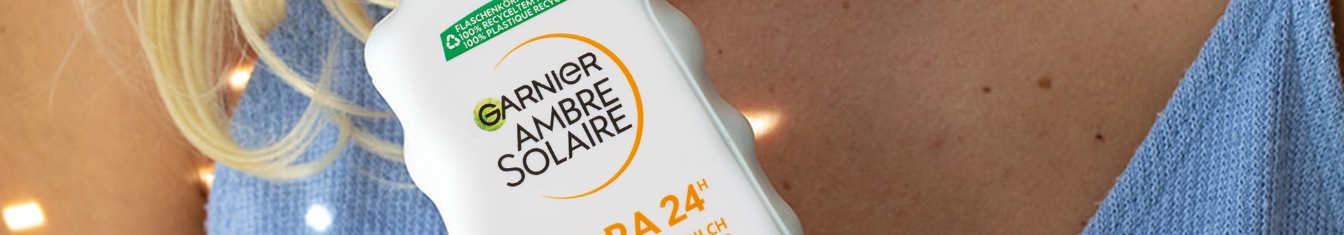 Blonde Frau, die die Garnier Ambre Solaire Hydra 24h protect+ Sonnenschutz-Milch LSF 30 in einer Hand vor ihrer Brust hält