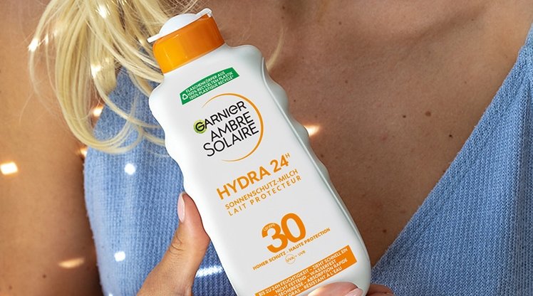 Blonde Frau, die die Garnier Ambre Solaire Hydra 24h protect+ Sonnenschutz-Milch LSF 30 in einer Hand vor ihrer Brust hält