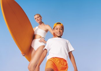 Ein kleiner Junge und eine Frau mit Surfboard vor blauem Himmel