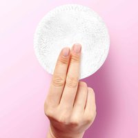 Eine Hand hält ein rundes, weißes Mikrofasertuch vor einen rosa Hintergrund.