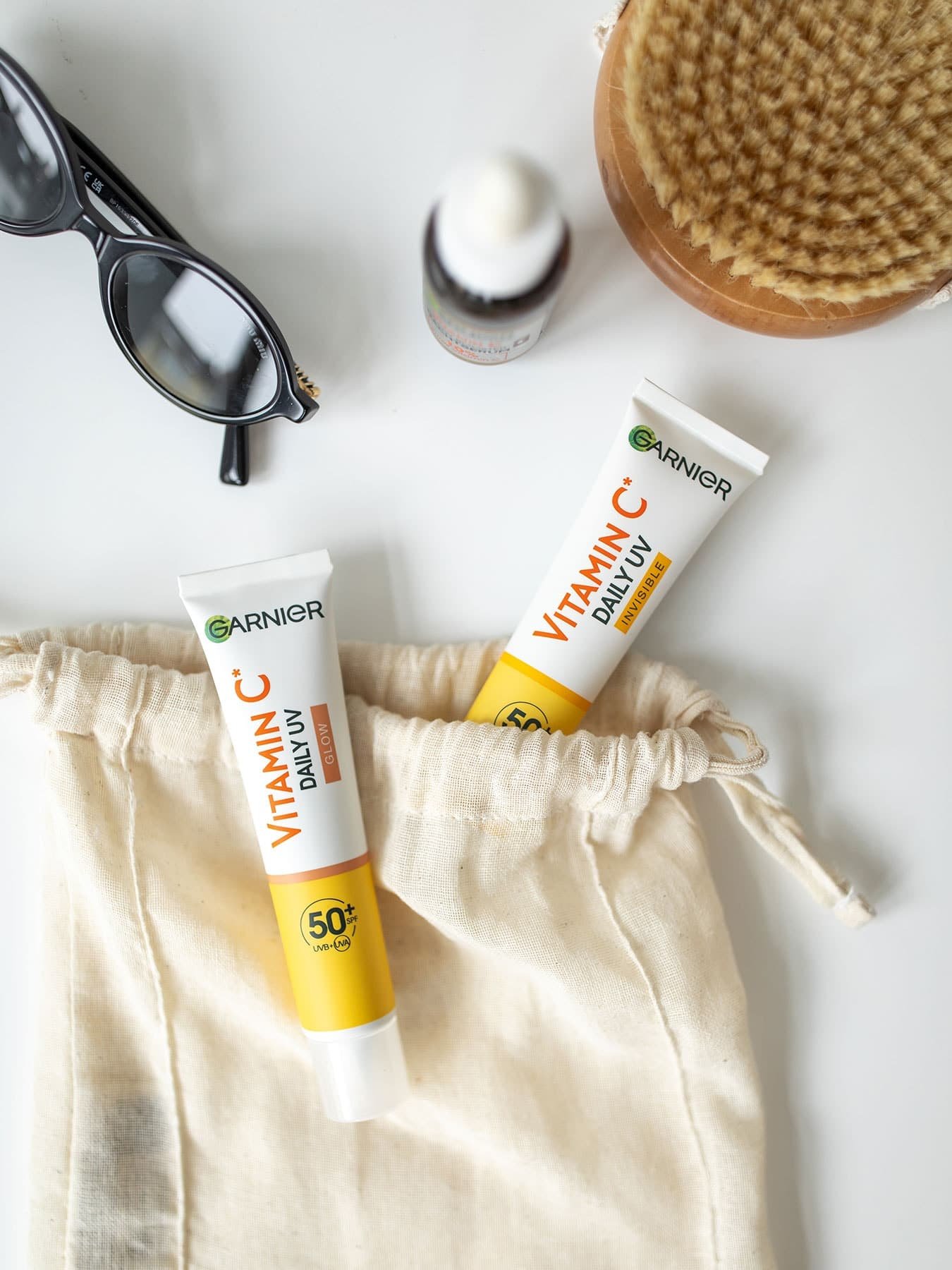 Stillleben mit Vitamin C Invisible & Super UV Sonnenfluid und einer Brille auf einer weißen Fläche
