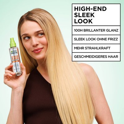 Ein junges, blondes, weibliches Model hält das Garnier Fructis Diamond Sleek Shine-Coat Glättigungsspray neben ihr Gesicht und schaut auf die Resultate - bis zu 100H brillianter Glanz, und strahles, extrem sleekes, geschmeidiges Haar.