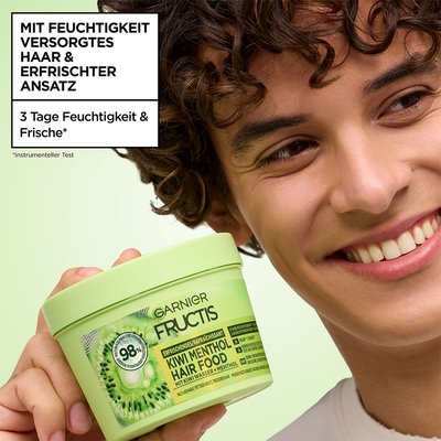 Mann mit lockigem Haar hält Fructis Multi-Use Haarmaske Kiwi Menthol Hair Food neben sein Gesicht und lächelt