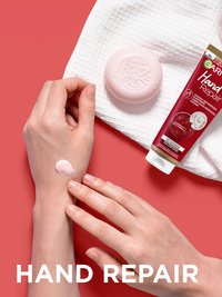 Hände einer Frau die etwas Creme auf dem linken Handrücken verteil und Produkt daneben liegend auf rotem Grund