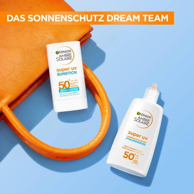 Alt Text: 2 Garnier Sonnenprodukte liegen auf einer orangenen Tasche, auf einem blauen Hintergrund. Der Garnier super uv sunstick LSF 50+ und das super uv Fluid mit Hyaluronsäure LSF 50+.