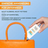 Der Garnier super uv sunstick wie er von einer Hand, vor einem blauen Hintergrund, in eine orangene Handtasche gesteckt wird.