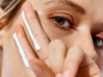 Nahaufnahme einer Frau, die in die Kamera blickt. Mit der linken Hand formt sie ein Peace-Zeichen; auf zwei ihrer Finger ist jeweils ein Cremestreifen.