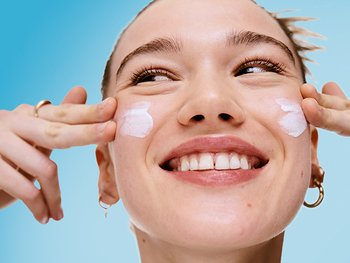 Eine junge Frau trägt mit ihren Händen Creme auf ihr Gesicht auf.