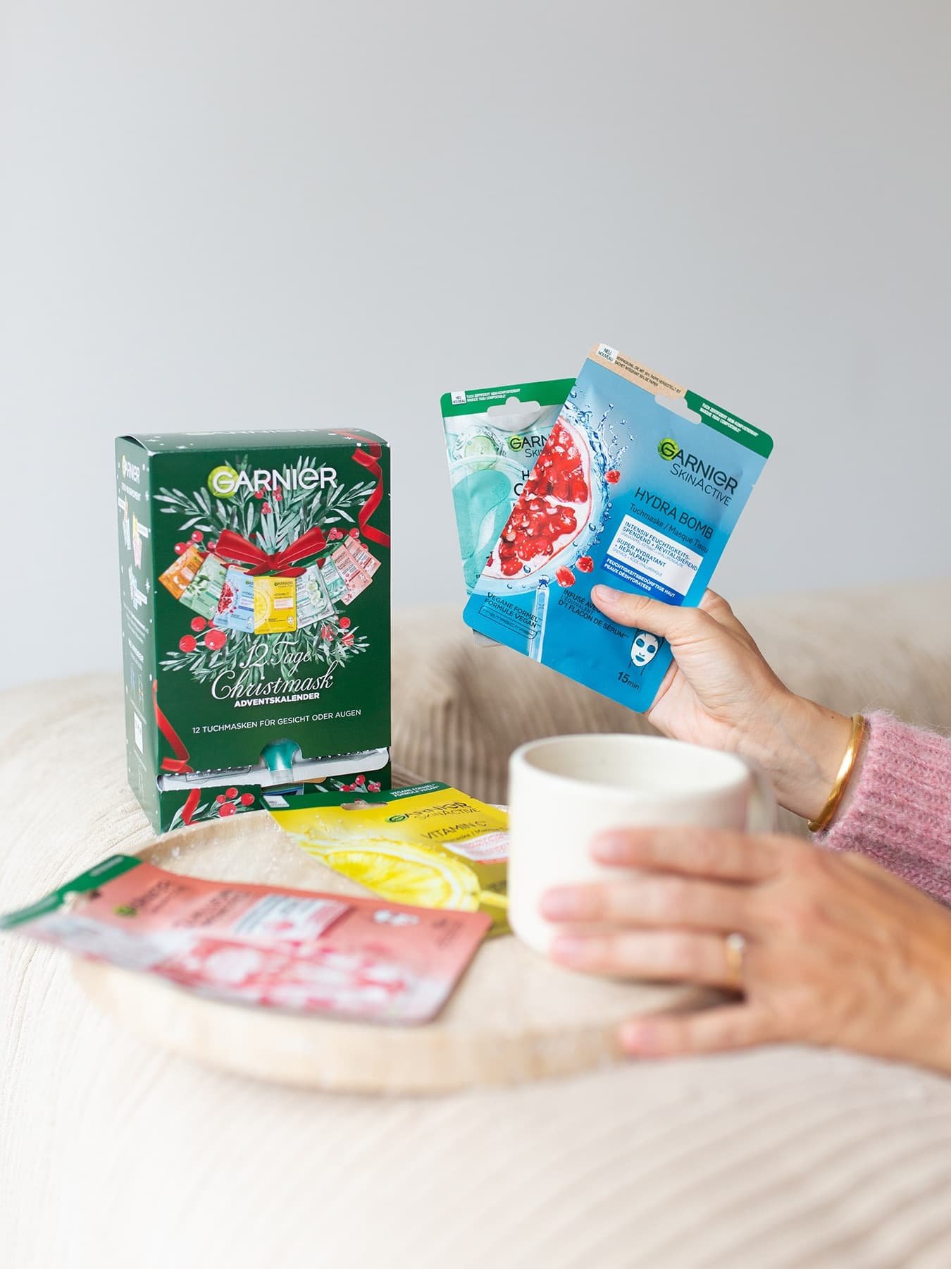 Tuchmasken Adventskalender teilweise ausgepackt und eine Frau mit Tasse in der Hand hält eine Tuchmasken-Verpackung in der anderen