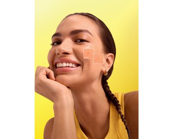 Lächelndes Model vor einem lebendigen gelben Hintergrund. Ihre Pigmentflecken werden aufgezeigt und der Vorher vs. Nachher Effekt des Garnier Vitamin C Glow Booster Serums darin aufgezeigt.